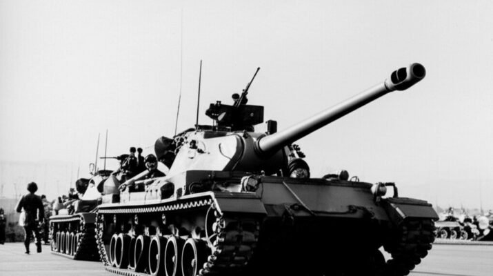 Barisan tank militer bergerak dalam parade resmi, menunjukkan kekuatan tempur dan dominasi militer di ruang publik.