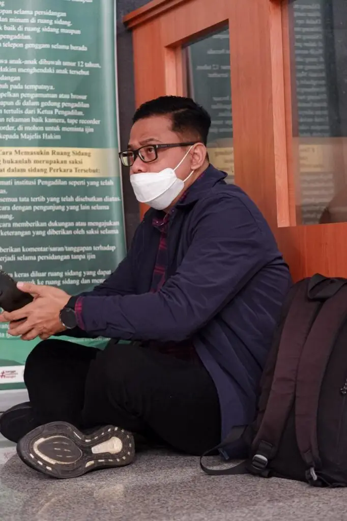 Tomy Ristanto duduk di lantai dengan masker wajah, memegang ponsel, di depan pintu ruang sidang, dengan tas ransel di sisinya, menunjukkan ketenangan di tengah menunggu hasil sidang Tom Lembong.