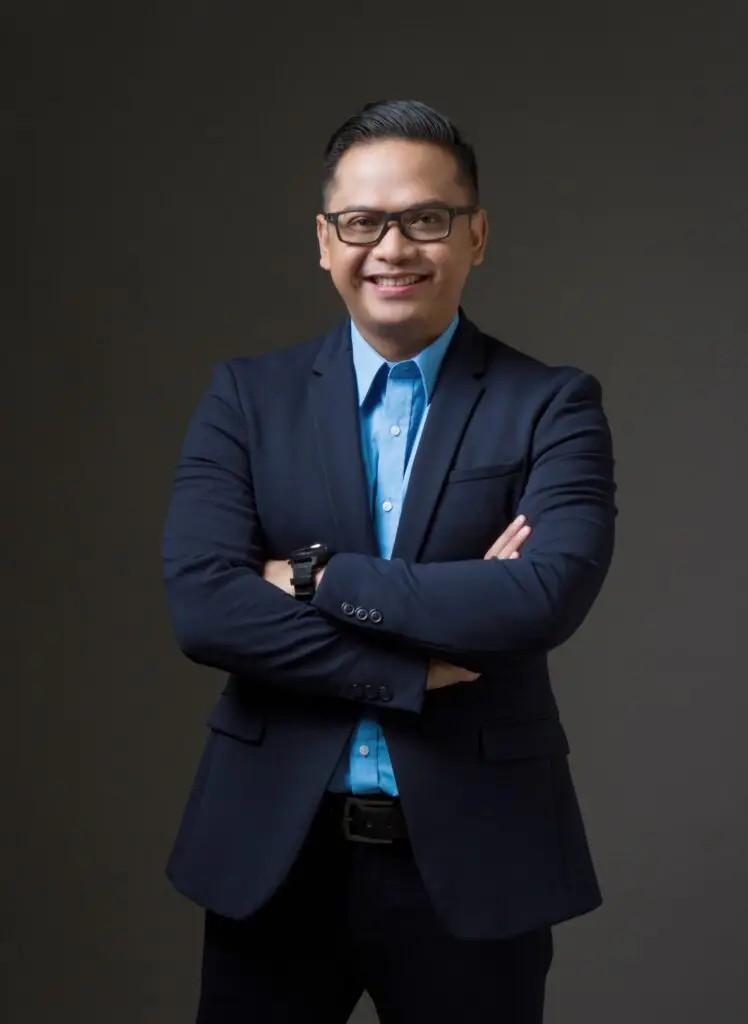 Tomy Ristanto berdiri dengan percaya diri, mengenakan setelan jas biru tua dan kemeja biru muda, dengan tangan bersilang, menunjukkan sikap profesional dan teguh